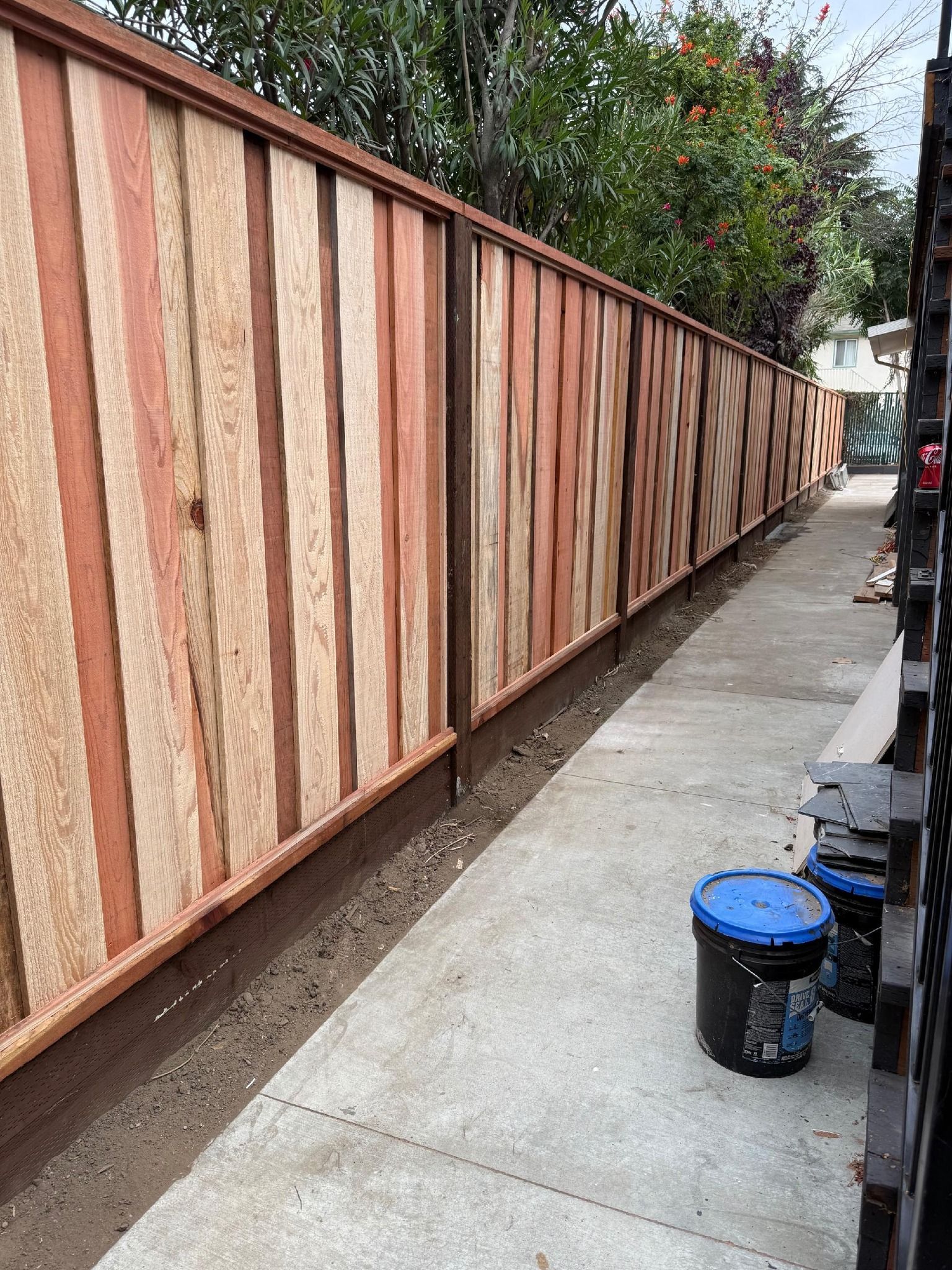 Long wooden fence with alternating light and dark panels along a concrete walkway, with paint buckets nearby.
