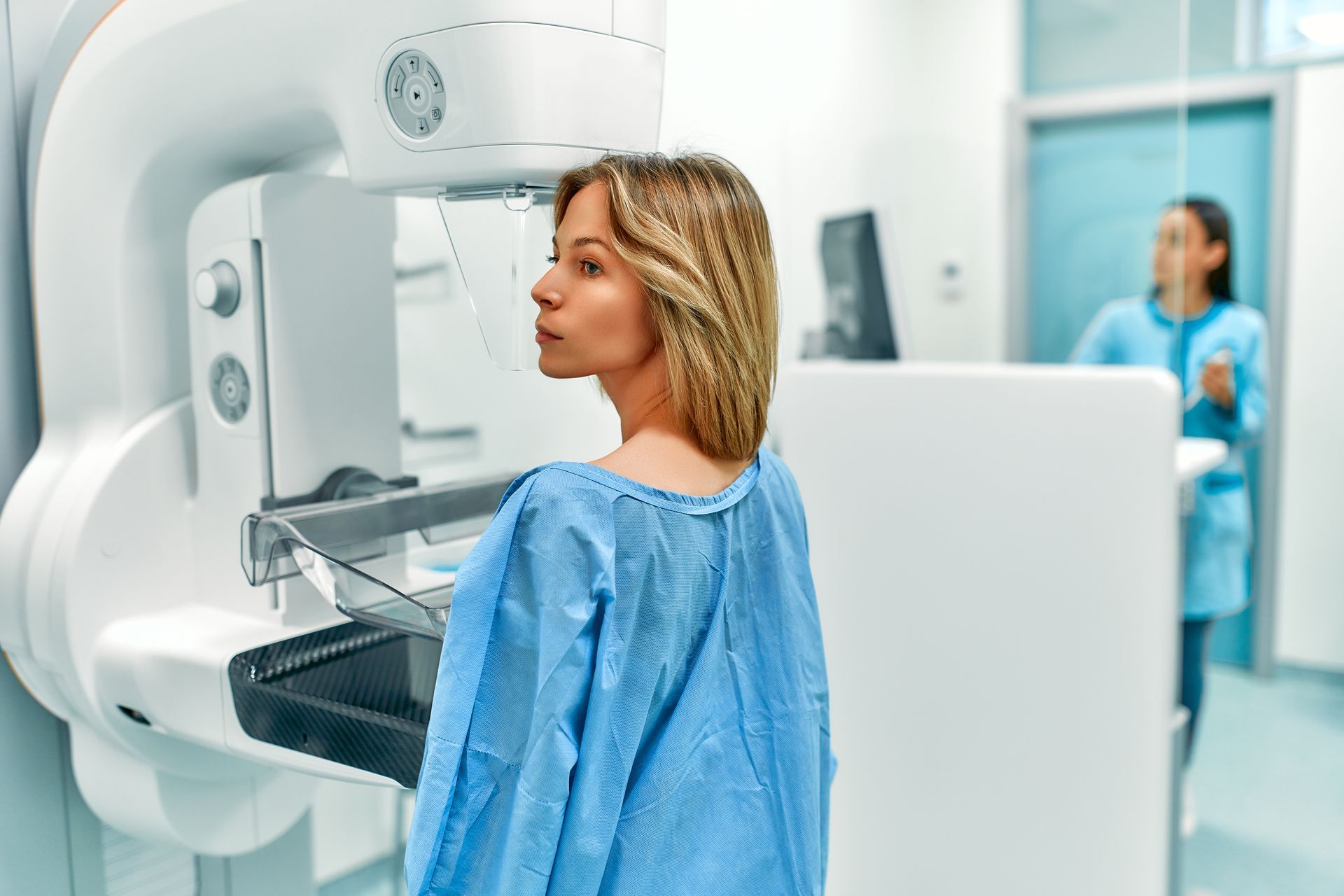 Patient in blue gown undergoing a mammogram in a bright clinic, with technician in background