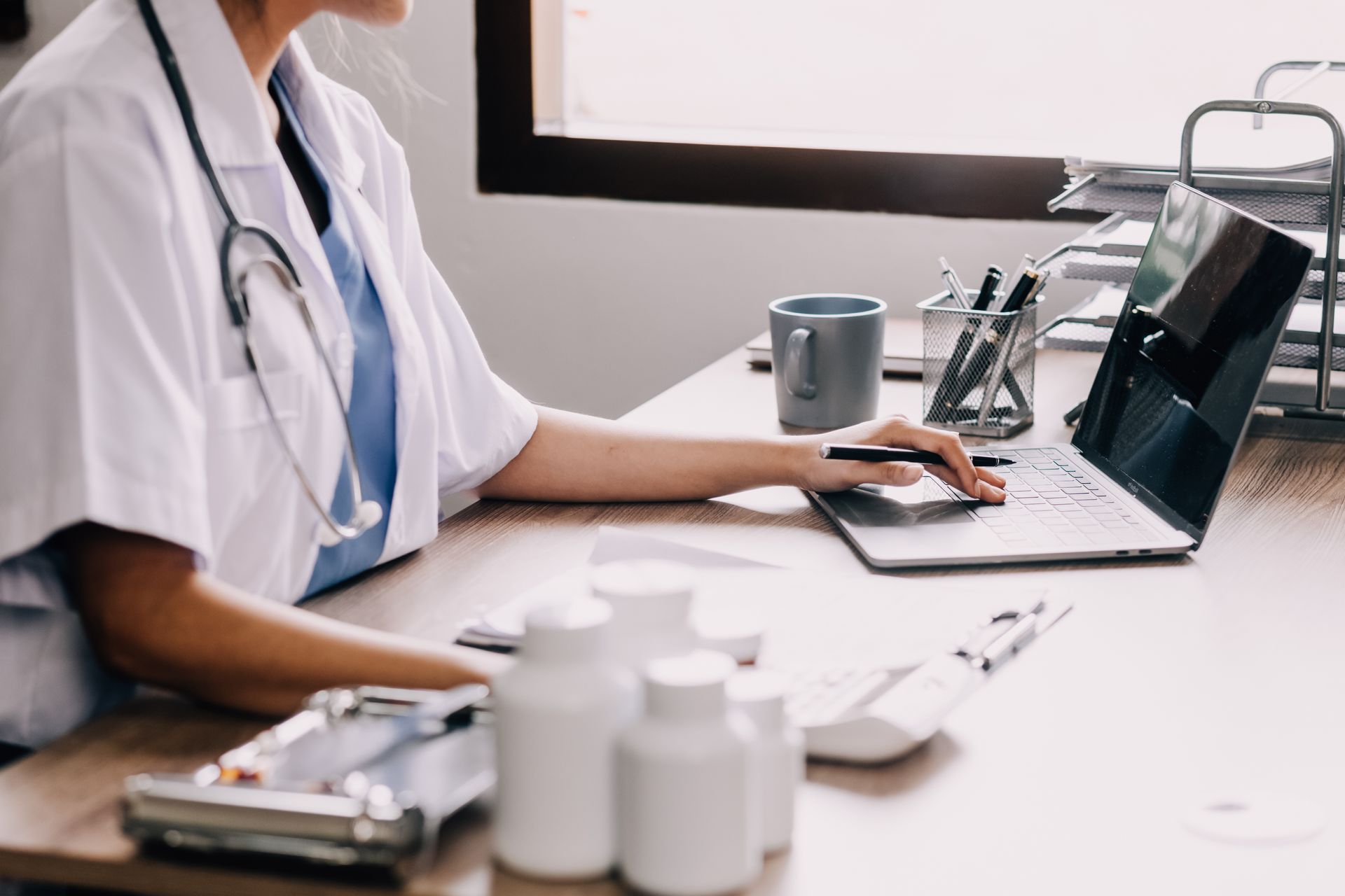 Doctor working on a laptop at a desk in a bright office, with a stethoscope and medical supplies nearby