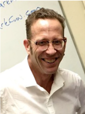 A man wearing glasses and a white shirt smiles in front of a white board
