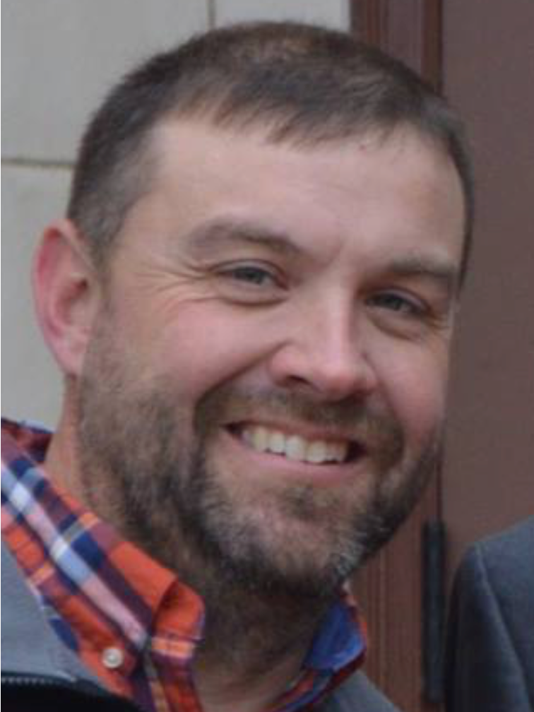 A man with a beard is smiling and wearing a plaid shirt.