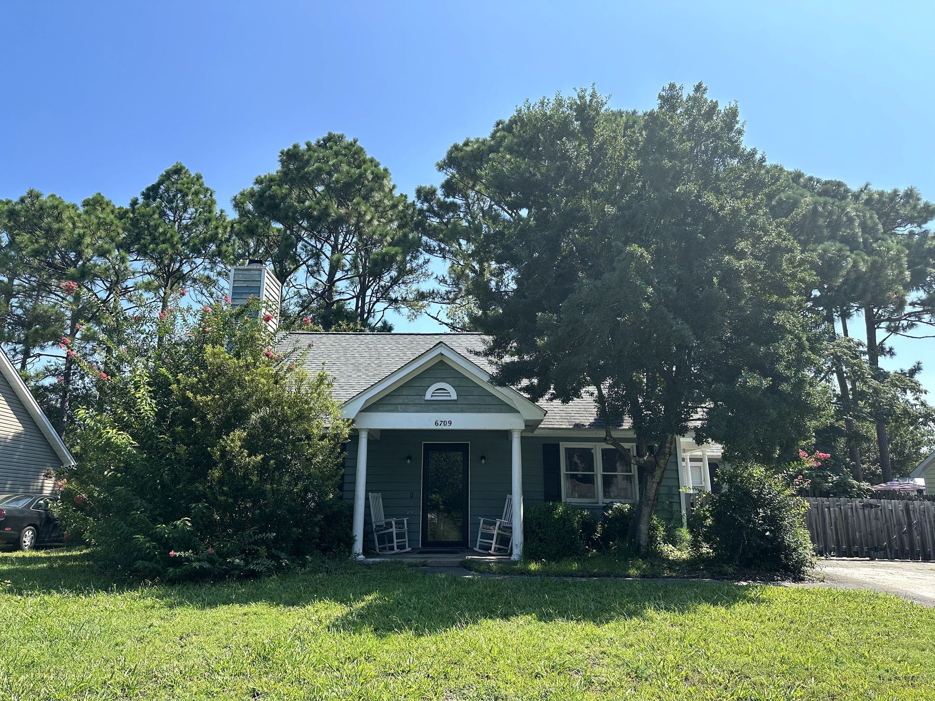 Small green house with a front porch and large trees, viewed from the front yard.