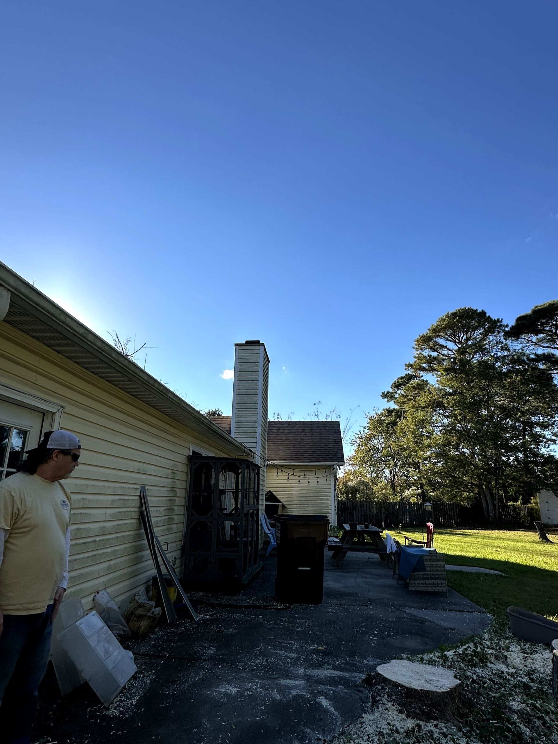 Person standing beside a white house and chimney in a sunny yard with trees and blue sky