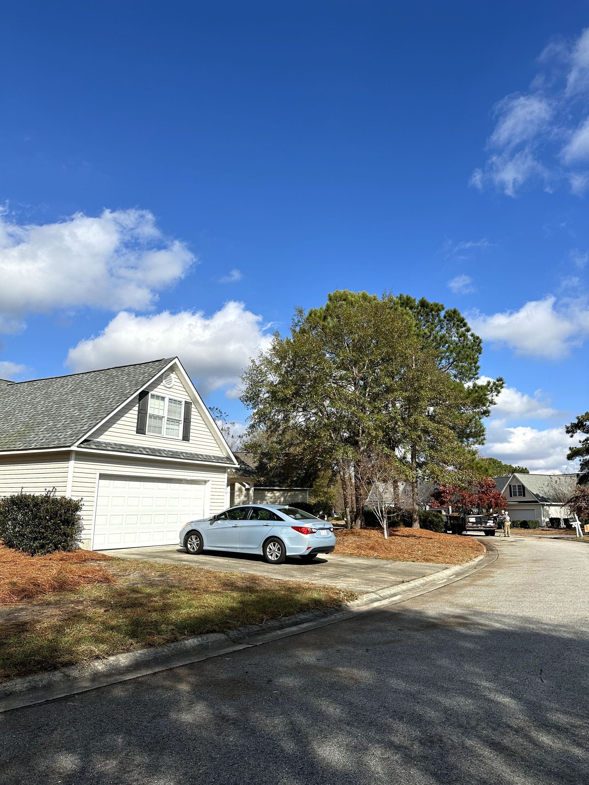 White house with a silver car, large tree, and gravel driveway under a blue sky with clouds