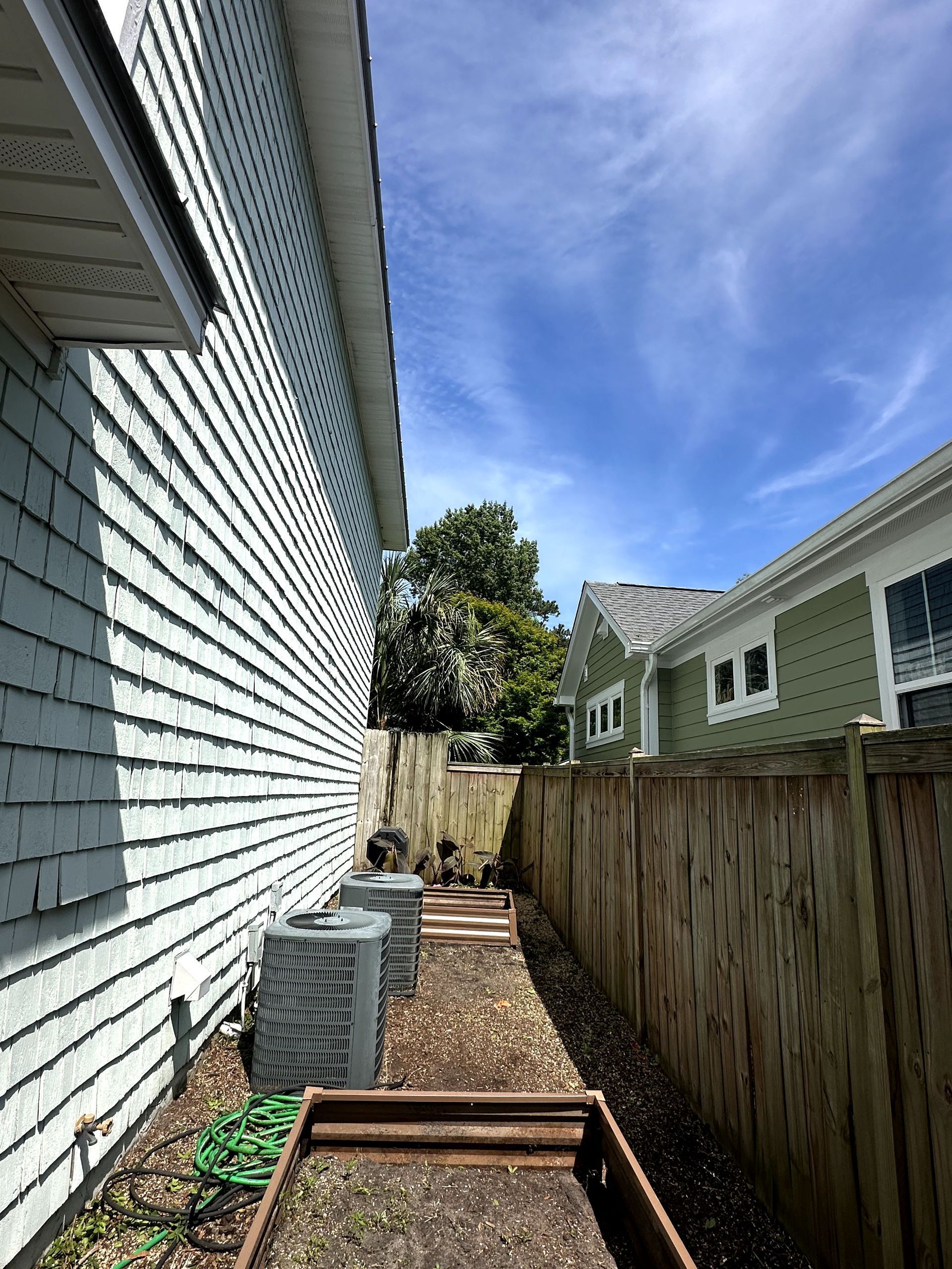 Narrow side yard between two houses with wood fence, gravel path, AC unit, and blue sky overhead