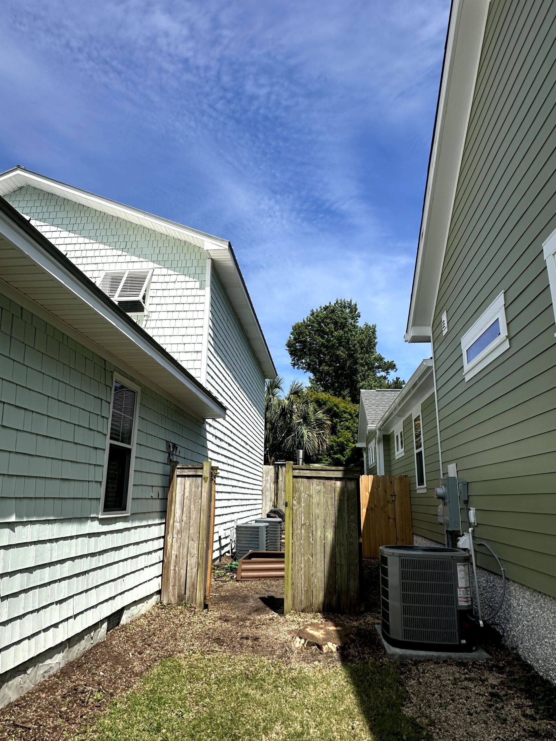 Narrow outdoor passage between two houses with gravel, fence, trees, and a clear blue sky.