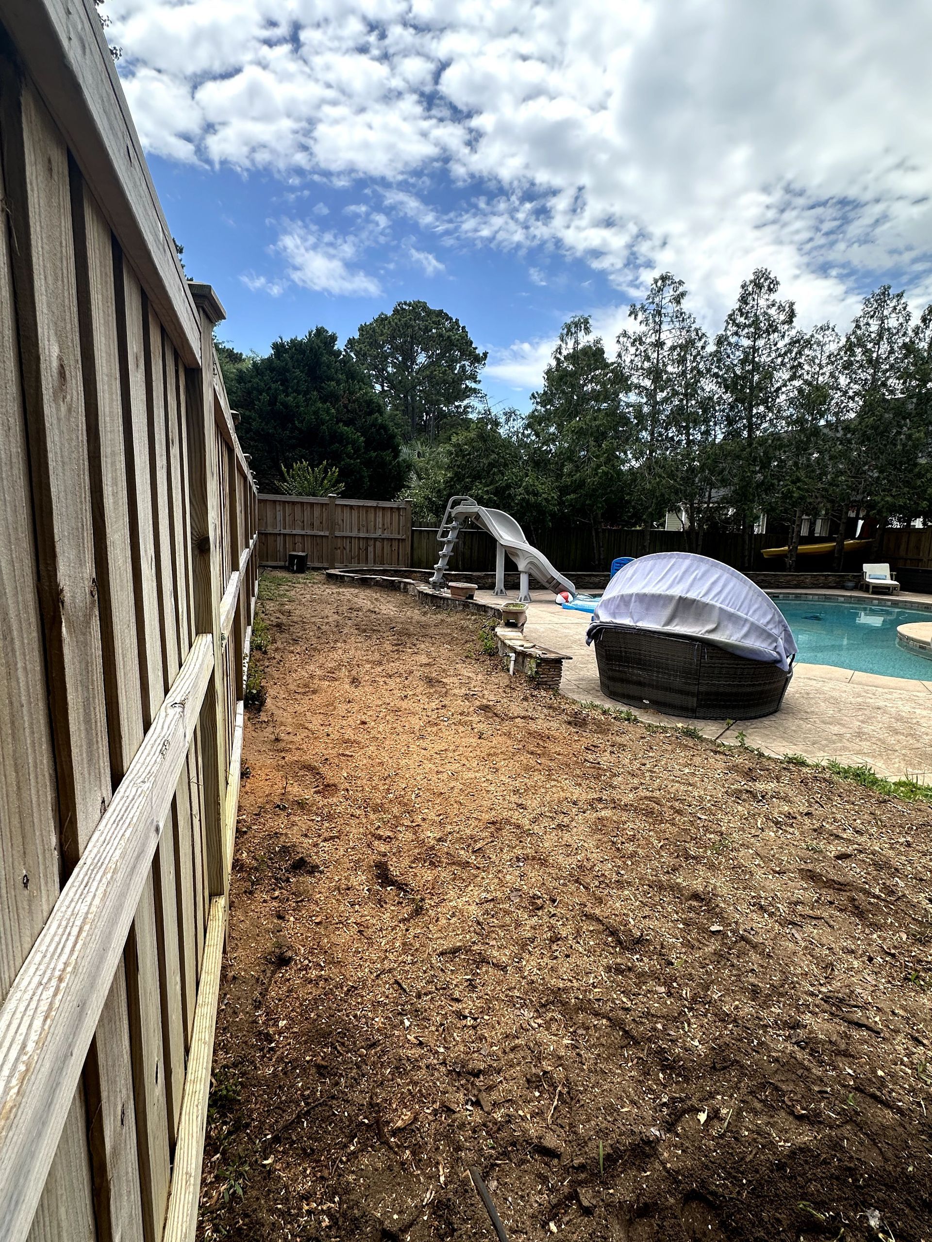 Backyard with wood chip ground, wooden fence, pool, and playground slide under a partly cloudy sky