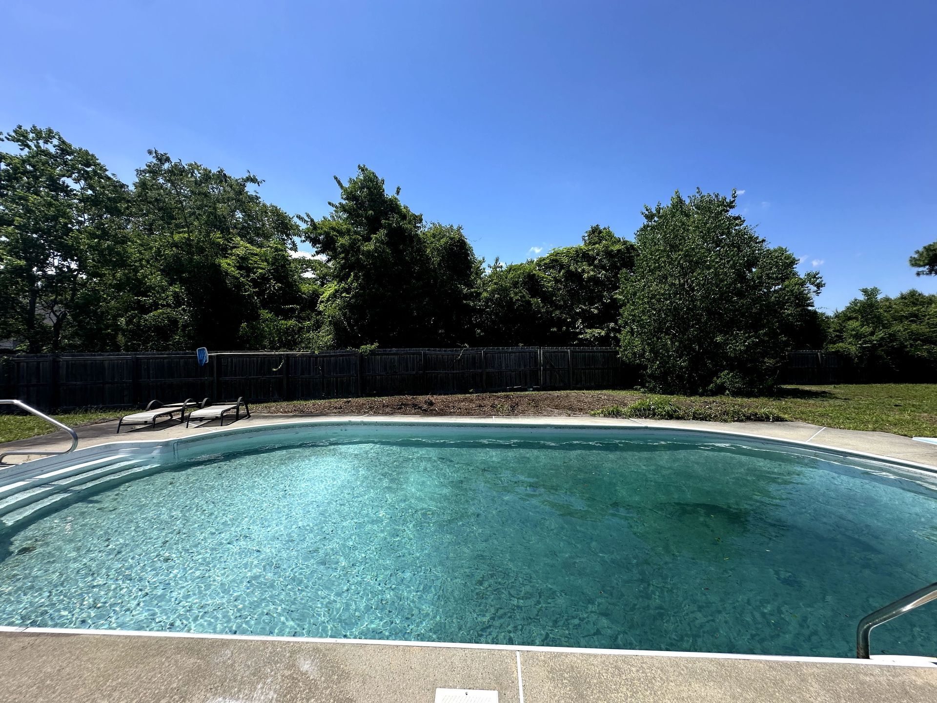 Backyard swimming pool with turquoise water, white deck, and trees under a clear blue sky