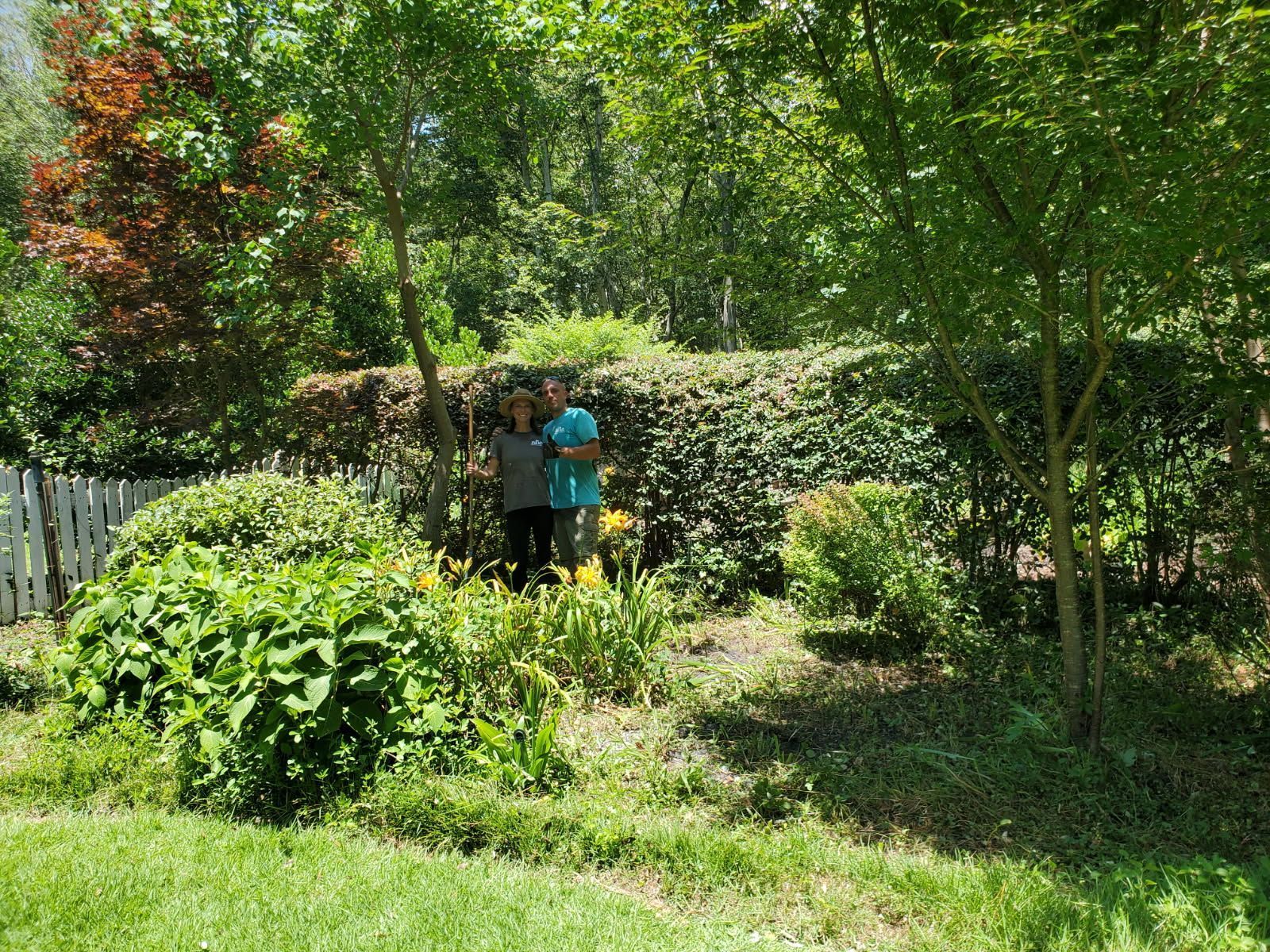 Person in teal shirt standing in a lush garden beside a hedge and trees