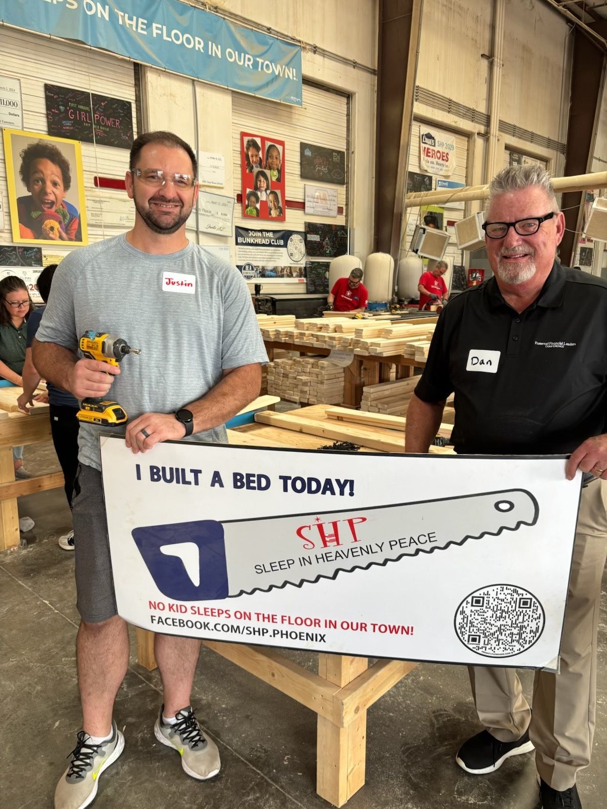 Two volunteers with tools and an I Built a Bed Today sign beside a bed frame