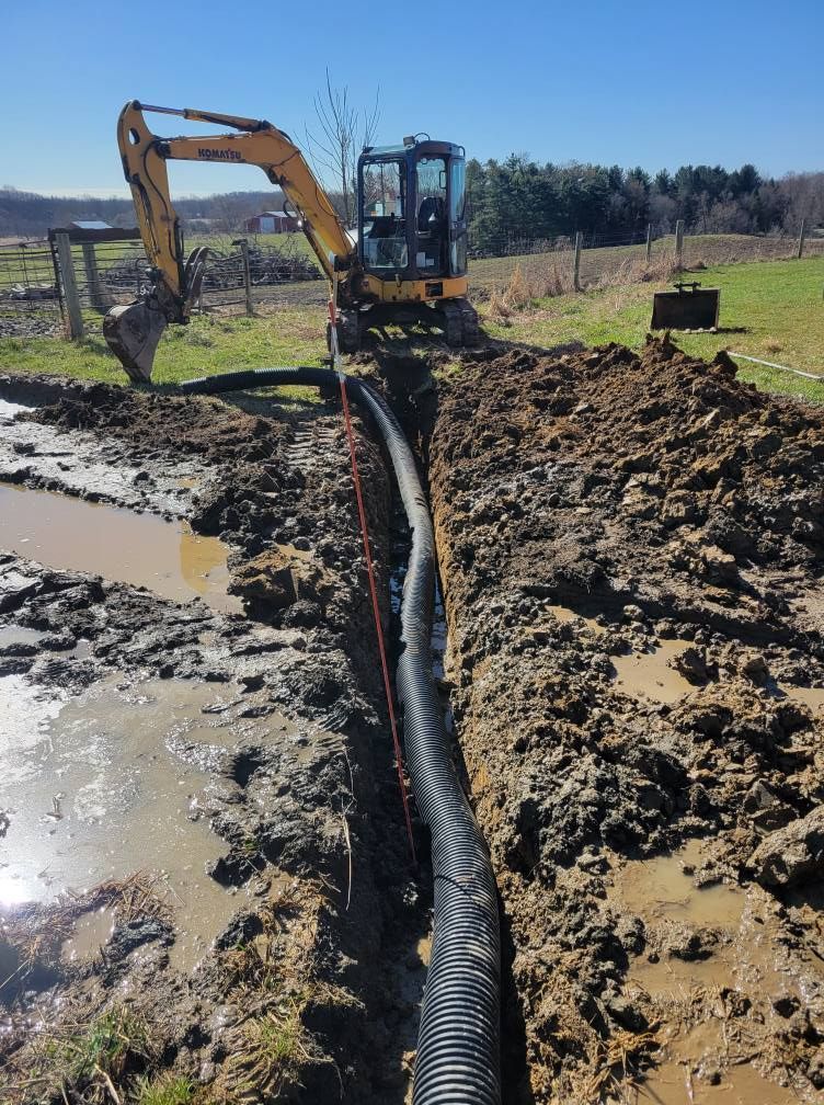 Excavator laying a black drainage pipe beside a muddy trench at a construction site