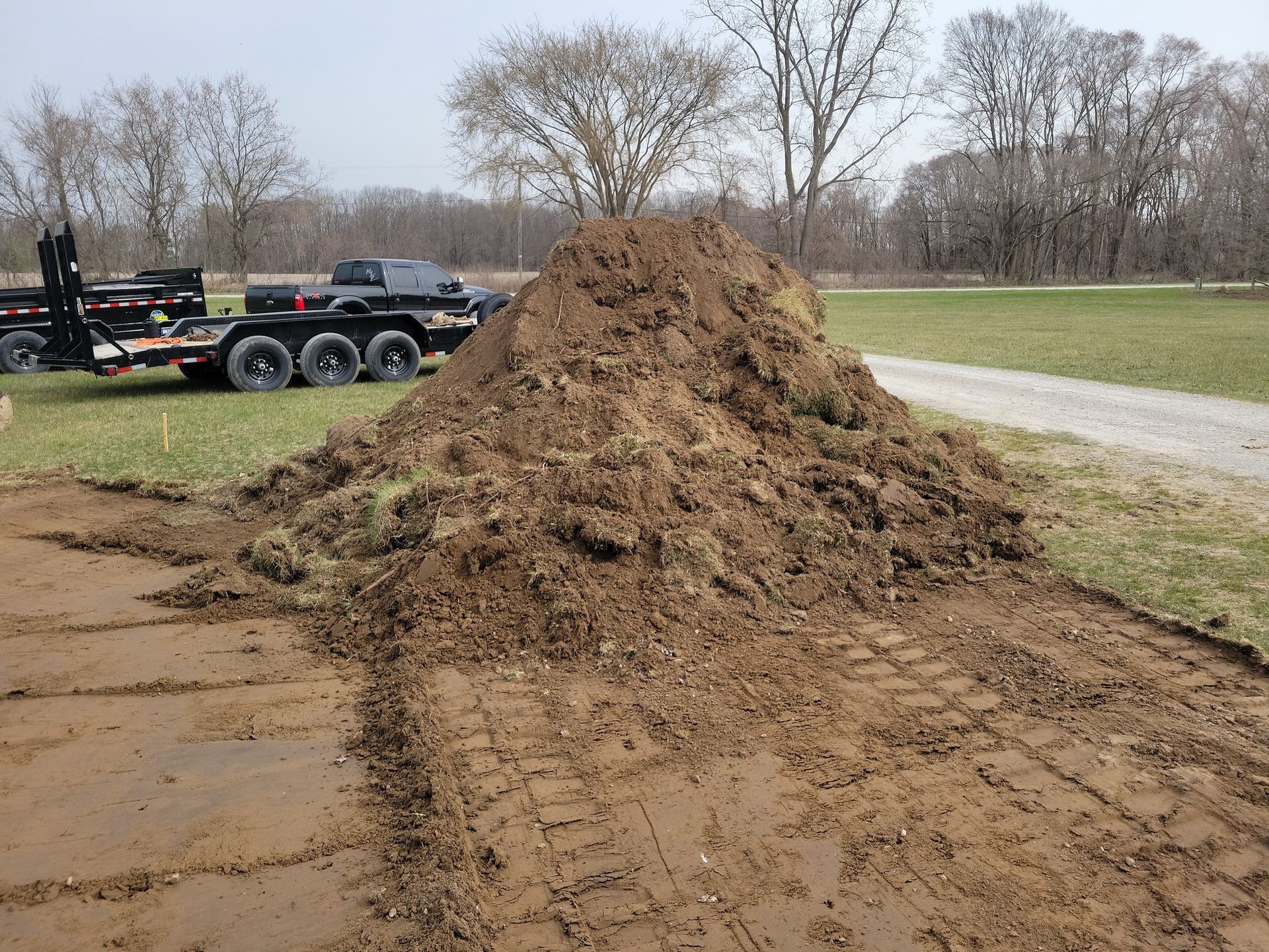 Large pile of dirt beside a road, with a trailer and bare trees in the background