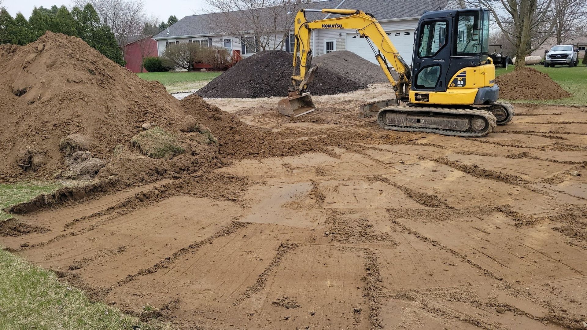 Yellow excavator on a muddy construction site beside piles of dirt and a house in the background