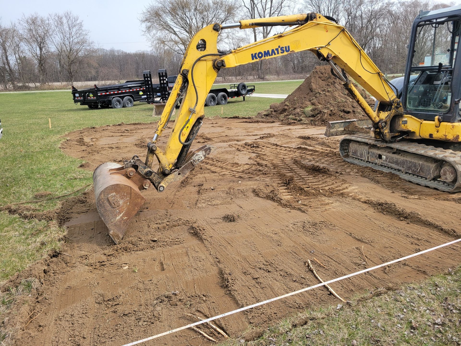 Yellow excavator grading dirt on a construction site near a grassy field and trees.