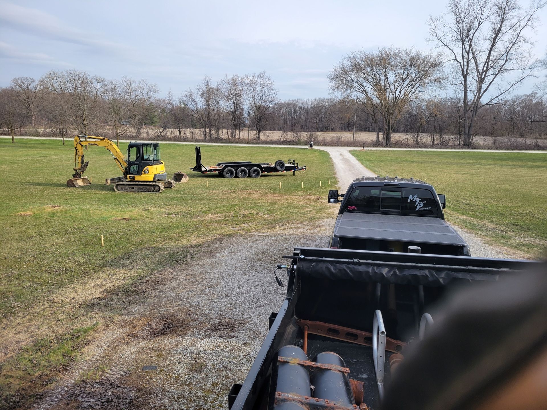 Backhoe and flatbed trailer on grassy field beside a gravel driveway, with trucks in the foreground and trees beyond