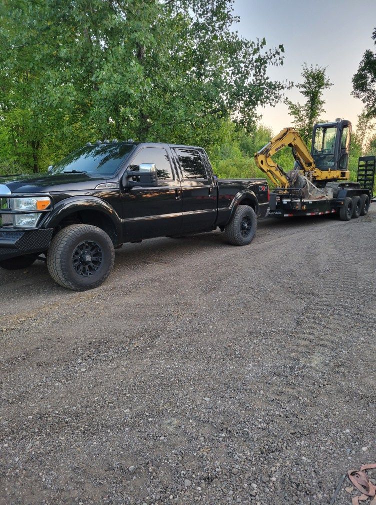 Black pickup truck parked on gravel, towing a backhoe trailer beside trees at dusk
