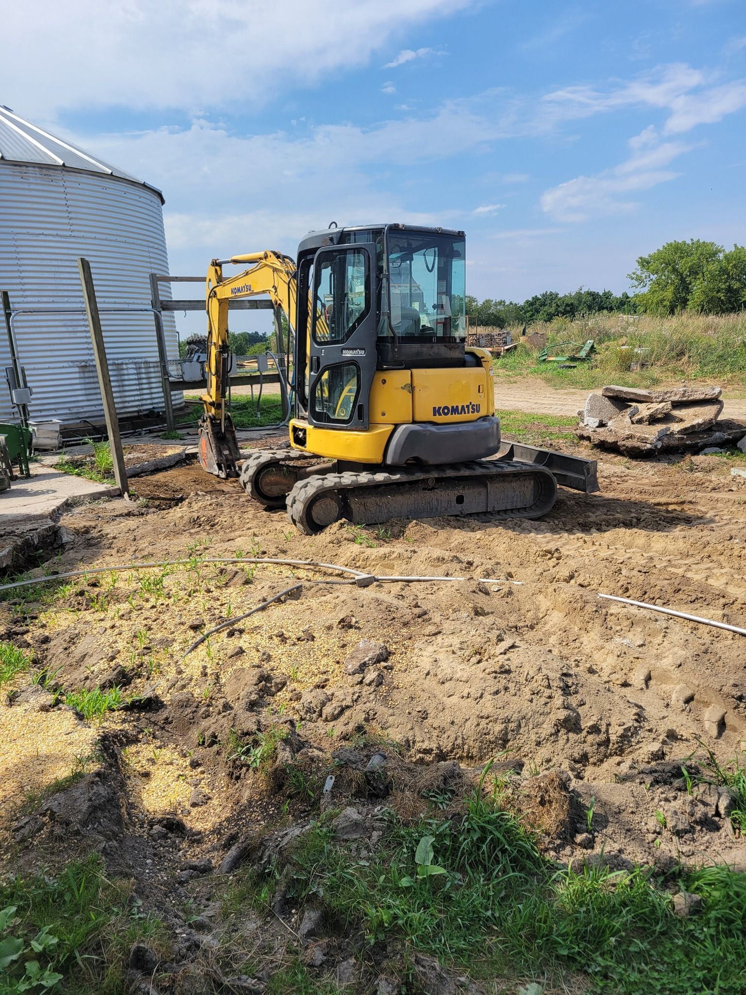 Yellow mini excavator on muddy ground beside a white building under a blue sky.