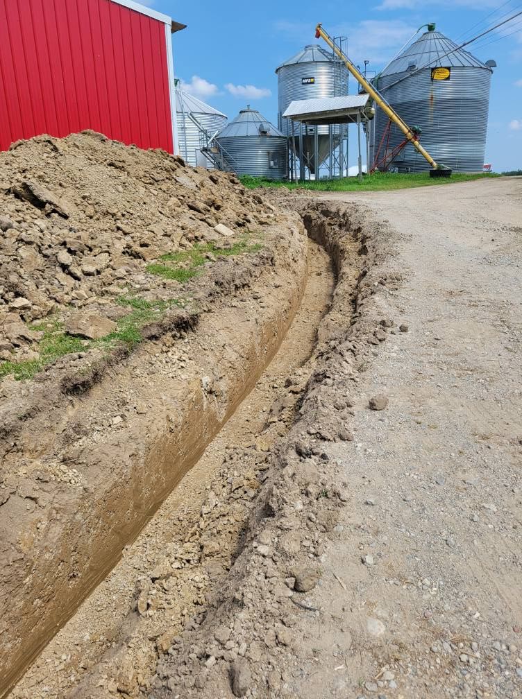 Freshly dug drainage trench beside a red barn on a gravel farm lane near grain silos