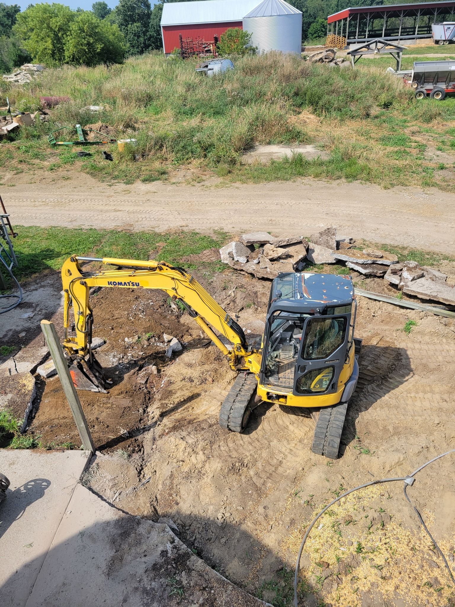 Yellow mini excavator on a dirt site with a barn and silo in the background.