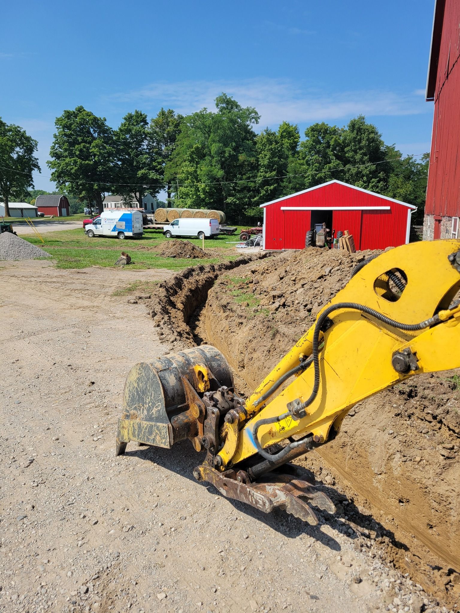 Yellow excavator digging a trench beside a red barn on a gravel construction site