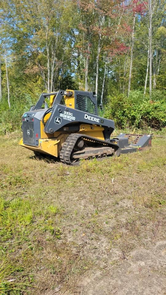 Yellow and black tracked skid steer on grass in a wooded clearing, with a front attachment raised.