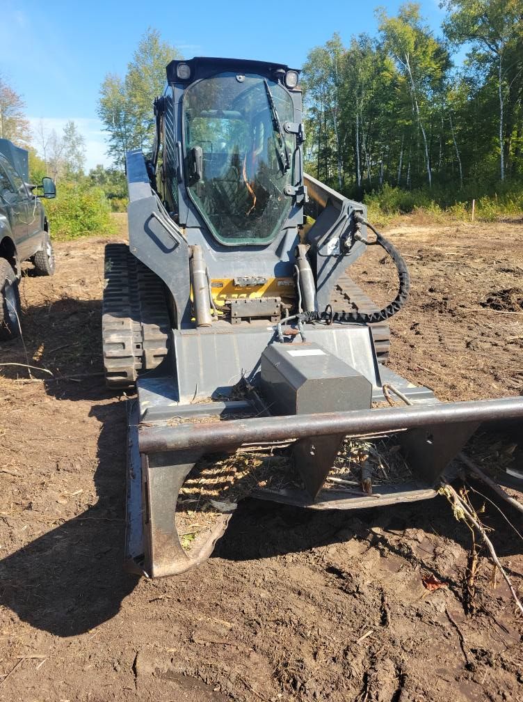Tracked skid-steer with brush cutter attachment clearing dirt on a muddy worksite in a wooded area