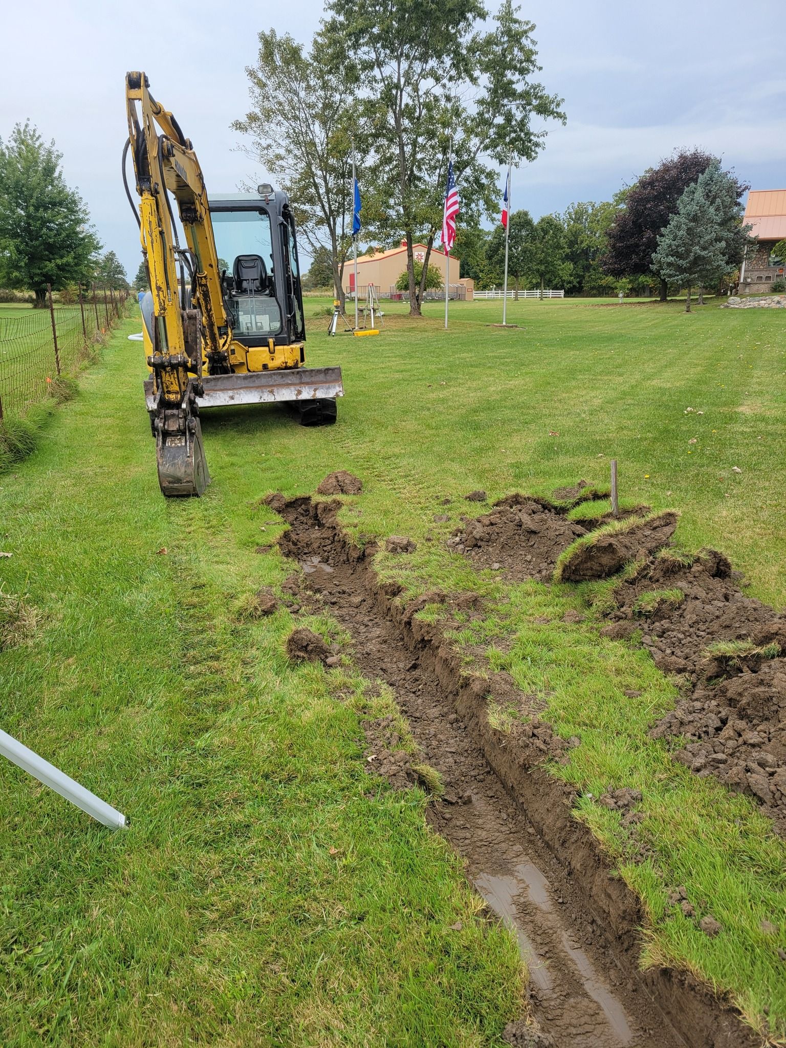 Small yellow excavator digging a trench in a grassy field near trees and a house