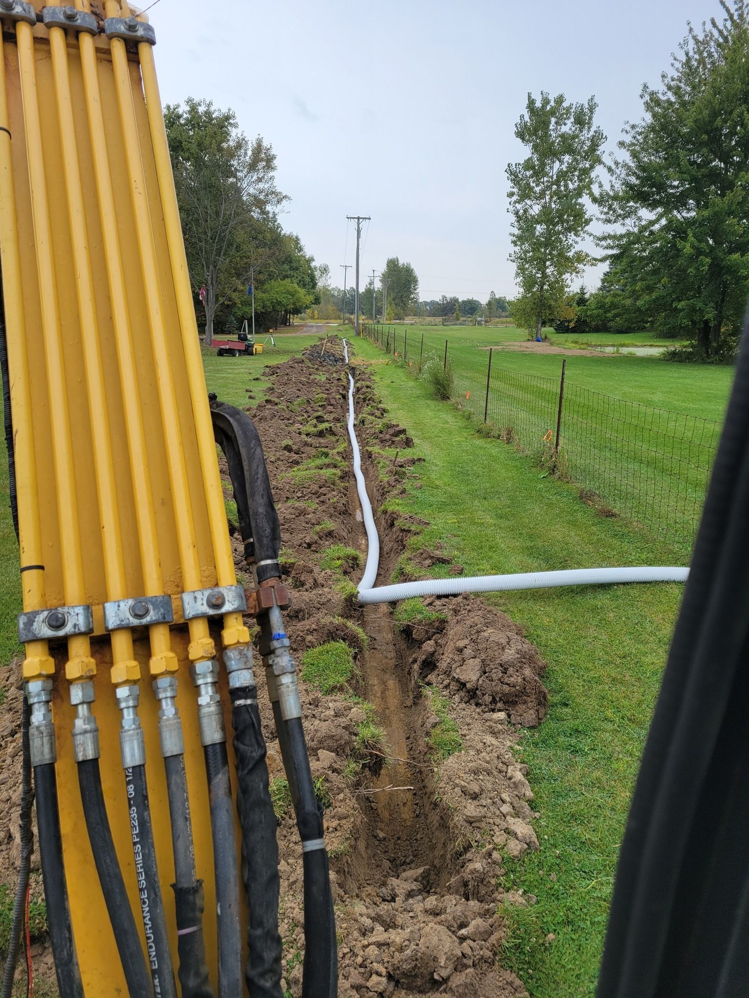 Yellow utility pipes beside a dug trench in a grassy rural field, with trees and a hose line