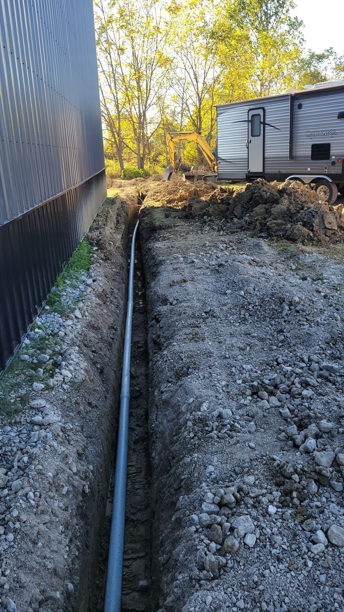 Gravel trench beside a building, with a black pipe running toward an RV in a wooded area.