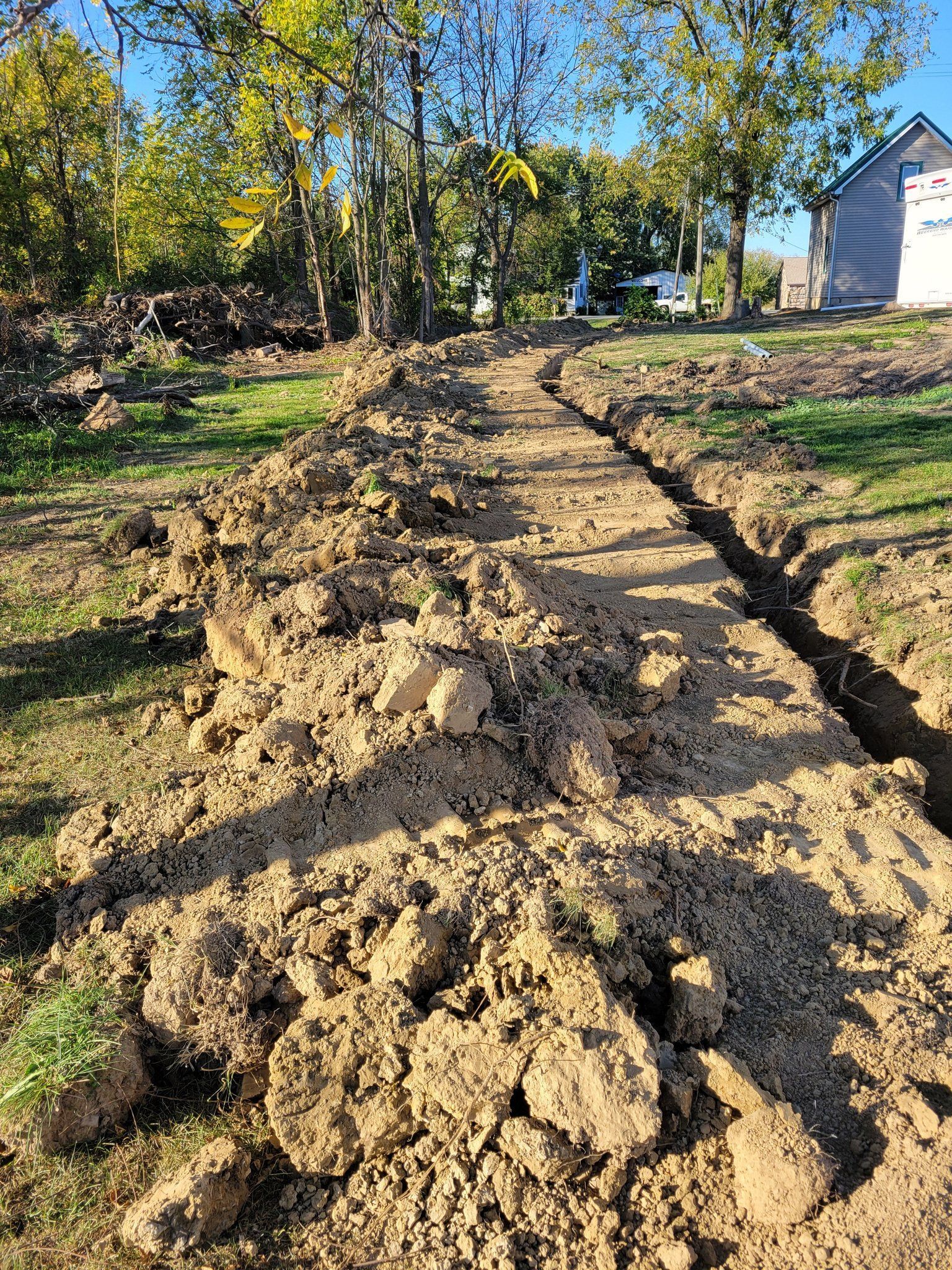 Freshly dug dirt path through a wooded yard beside a white house on a sunny day