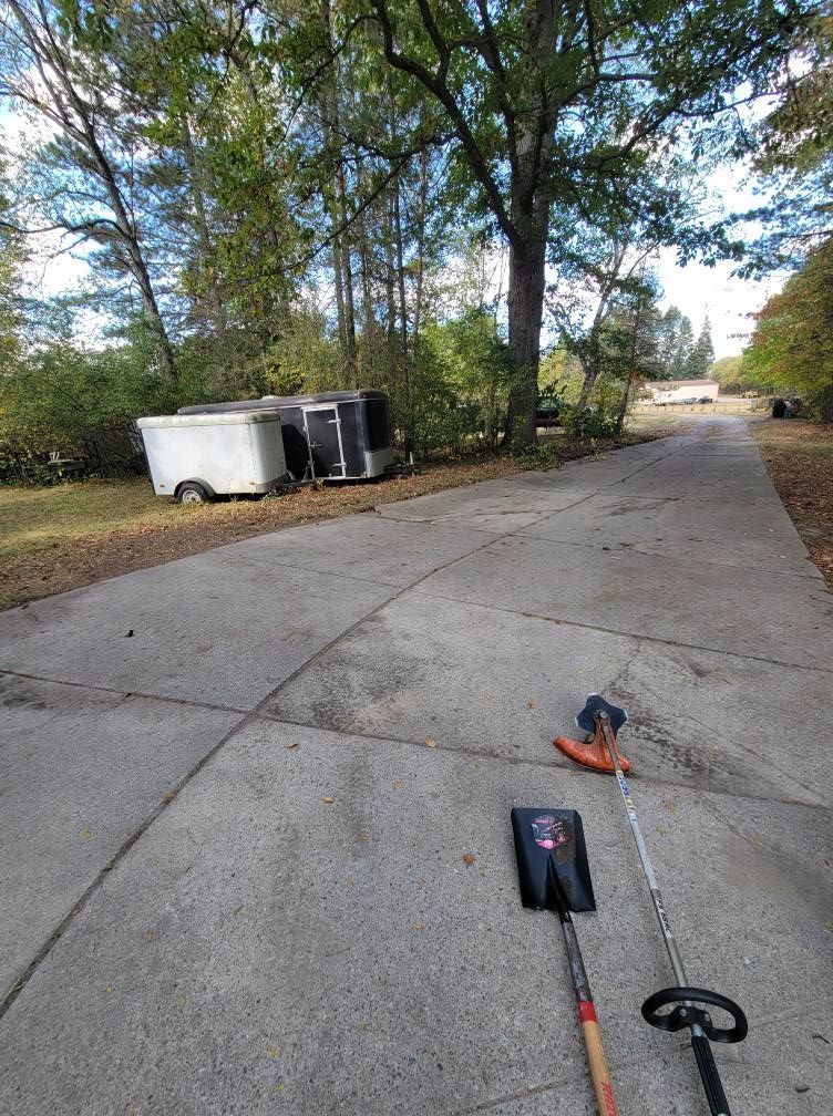 Concrete driveway with scattered yard tools, trees, and a trailer in the background