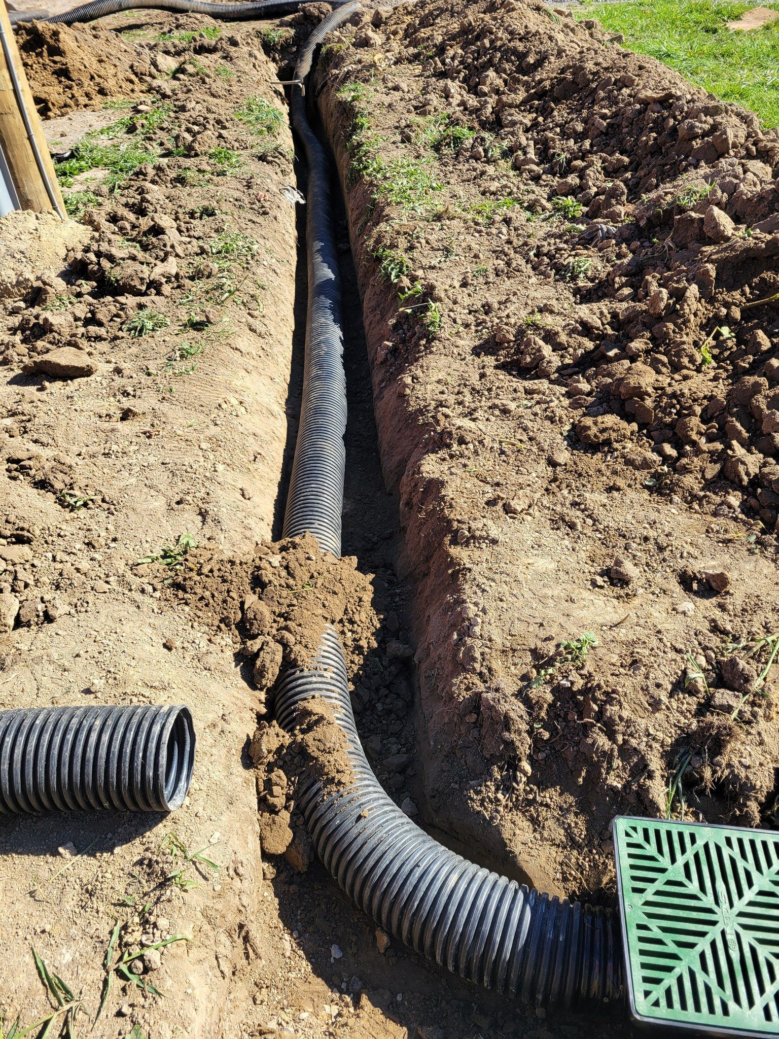 Black corrugated drainage pipe laid in a dirt trench outdoors, with a curved section at the end