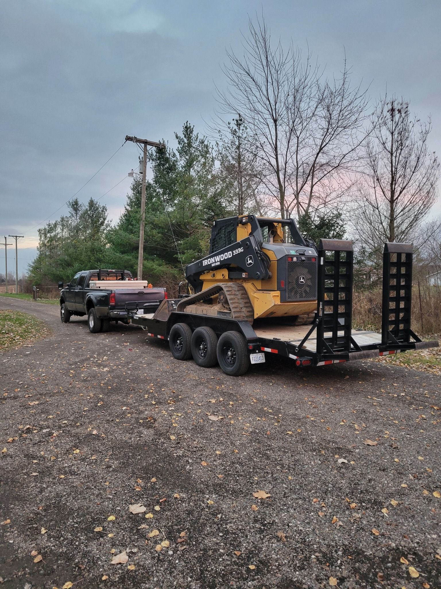 Truck towing a trailer with a skid steer loader on a gravel road at dusk