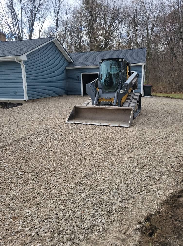 Front loader parked on a gravel driveway beside a blue house, with leafless trees in the background