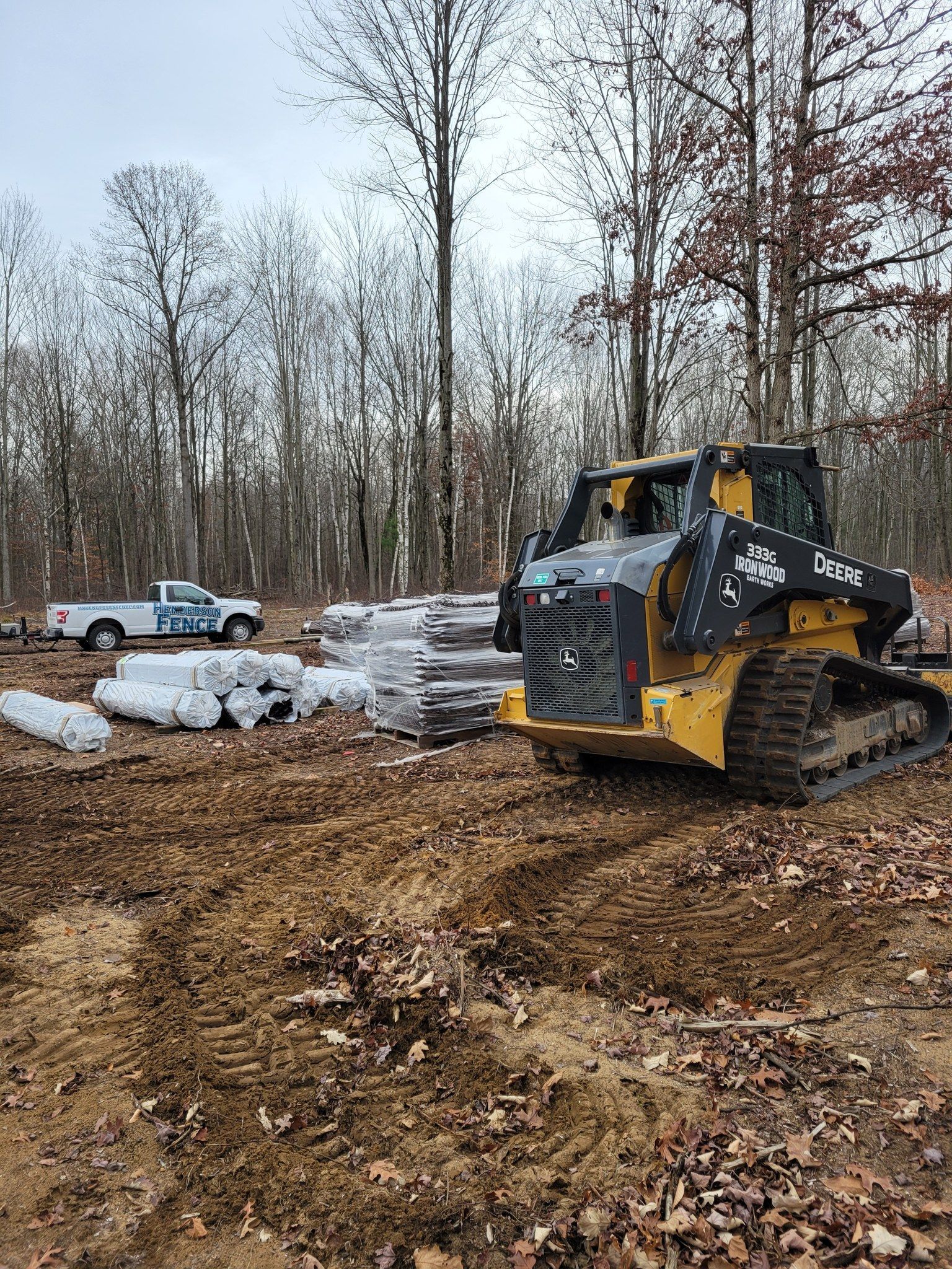 Yellow skid steer on muddy clearing with snow piles and bare trees in the background