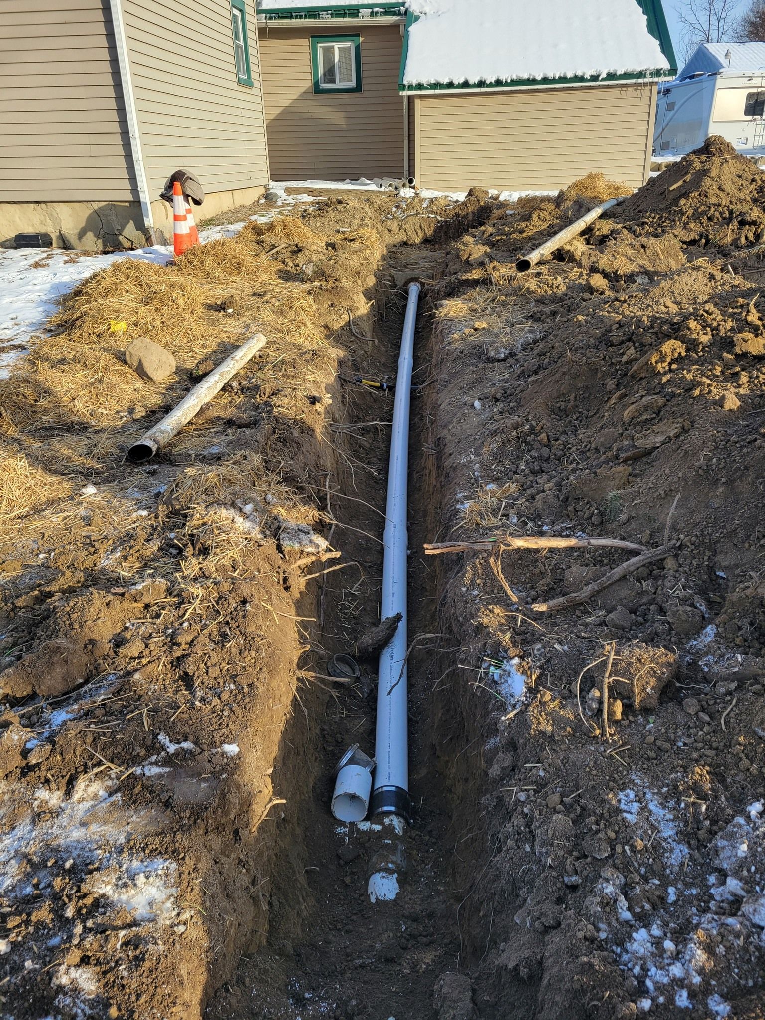 Trench with a white pipe running through dirt beside a house, with a worker in the background.