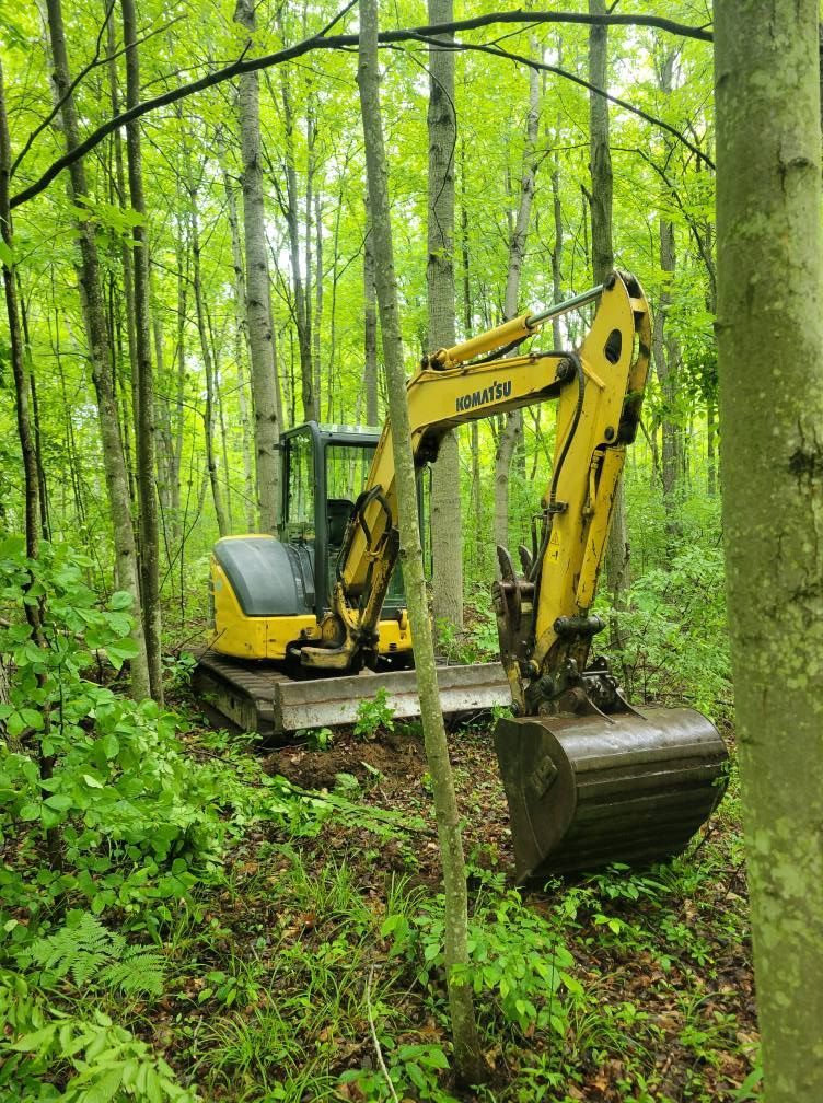 Yellow excavator in a green forest, moving a large log on muddy ground.