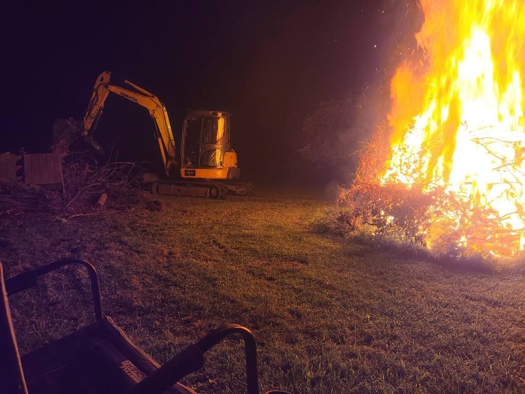 Excavator beside a large blaze at night, with flames lighting a dark field.