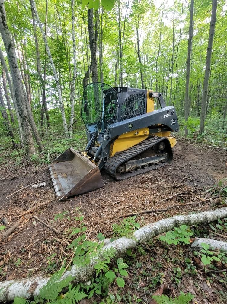 Black-and-yellow skid steer with bucket on a forest trail, clearing dirt and debris among trees.