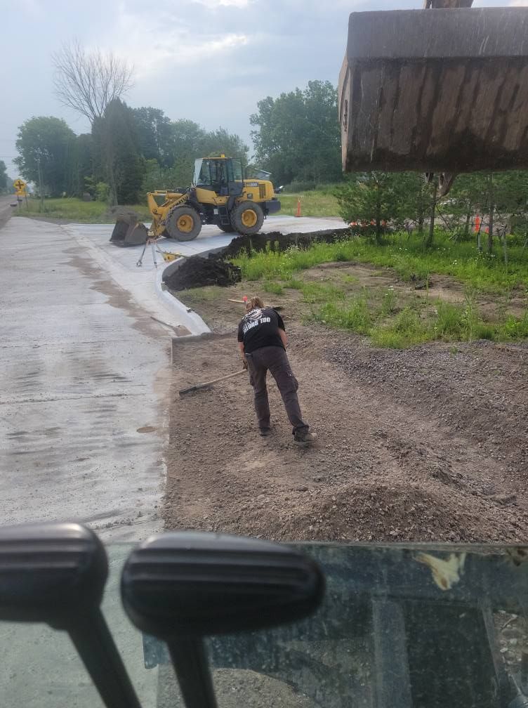 Worker standing on gravel beside a road construction site with a yellow loader and wet pavement