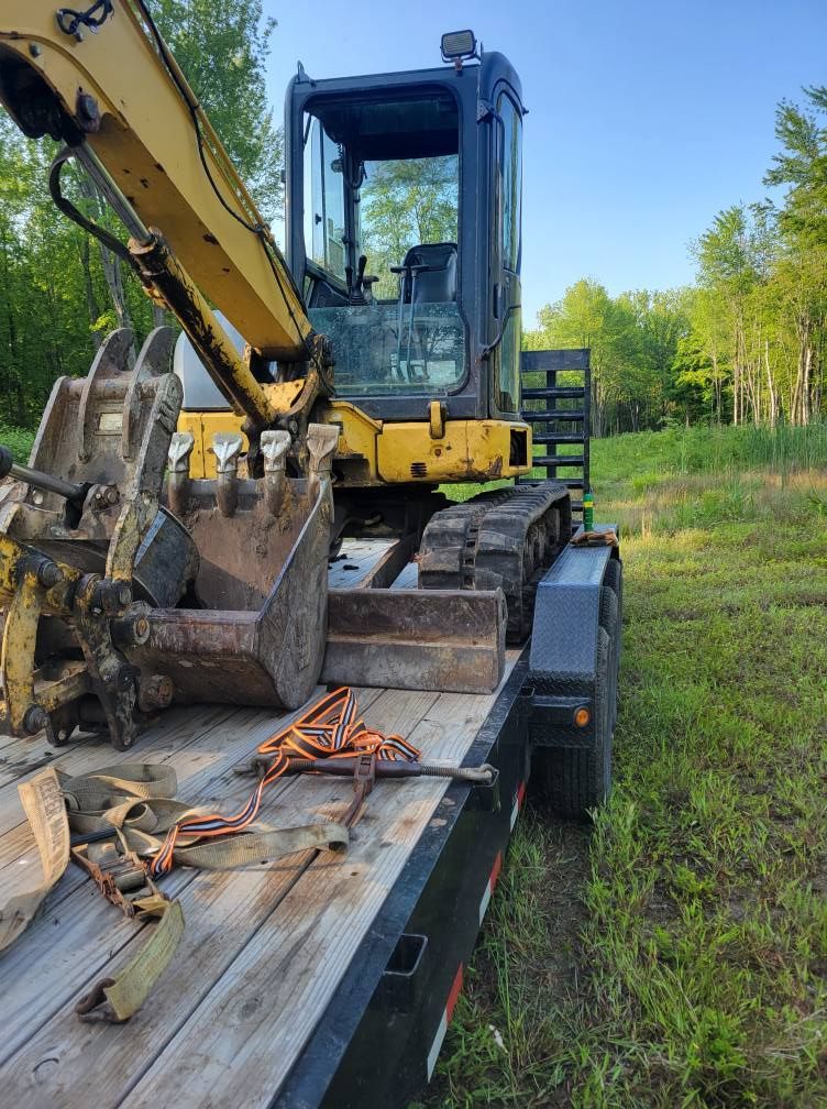 Yellow excavator on a flatbed trailer in a grassy field, with orange straps securing it.