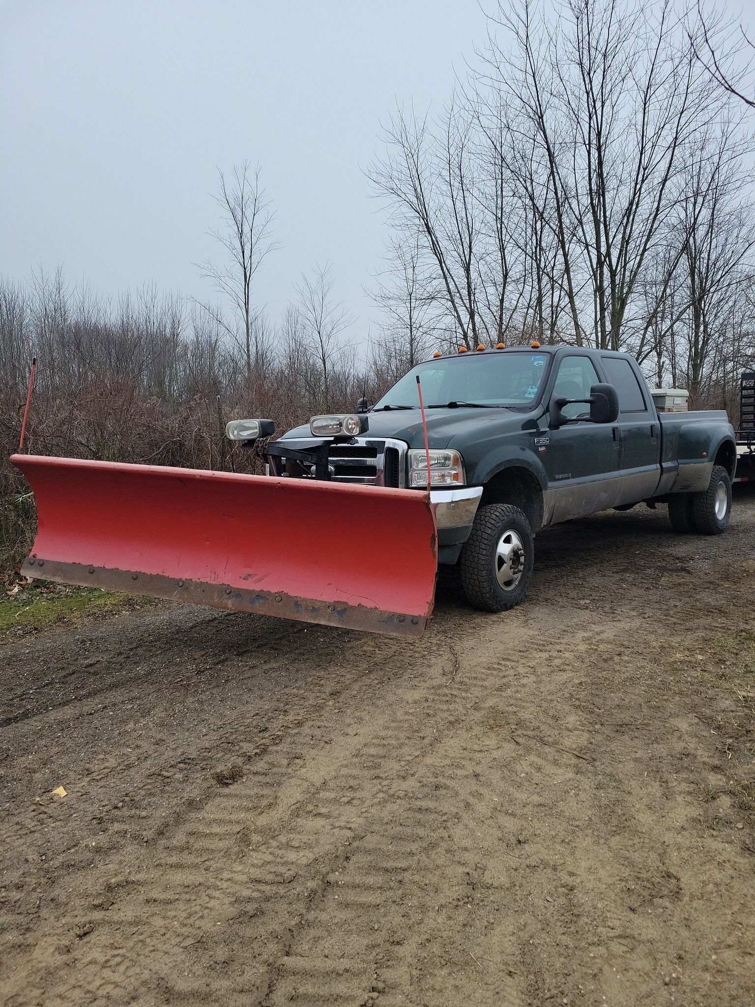Dark pickup truck with a red snowplow blade parked on a muddy gravel road.