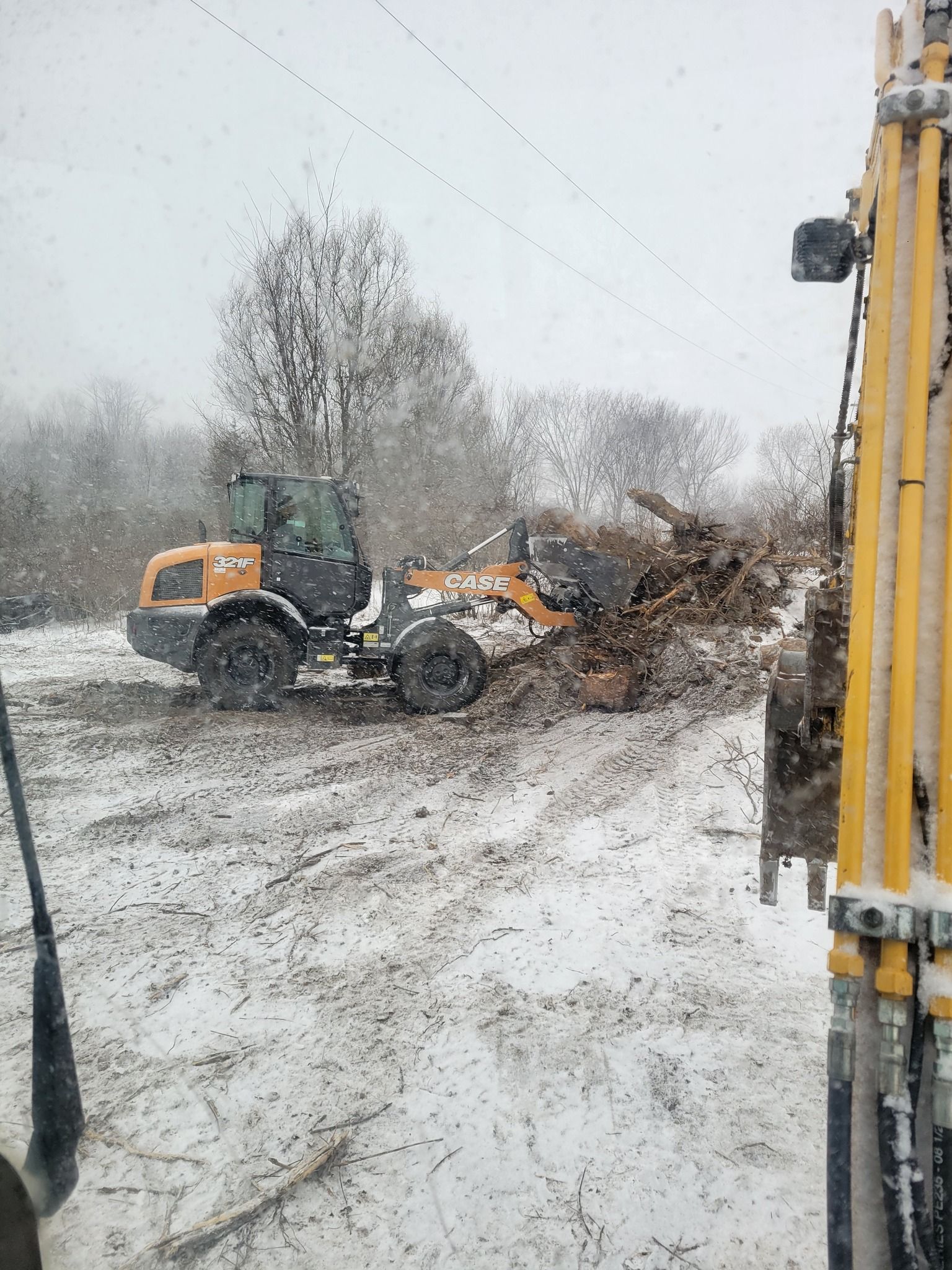 Orange skid steer pushing a large pile of branches in a snowy yard beside a yellow truck