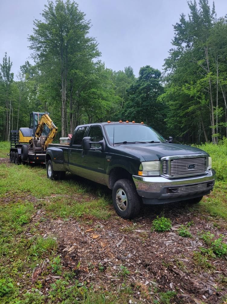 Black pickup truck towing a trailer with a yellow excavator on a wooded dirt path