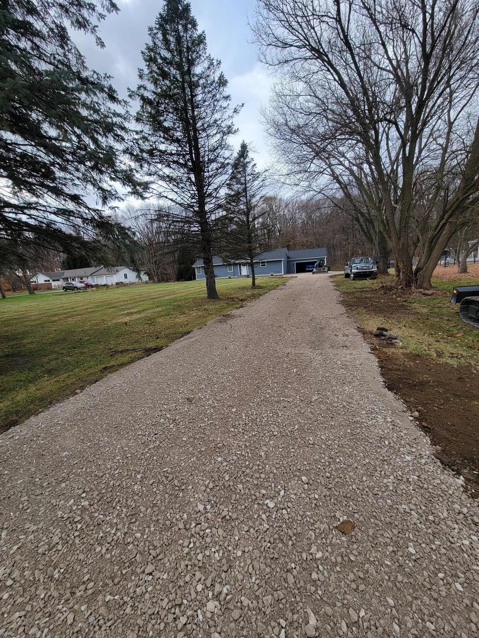 Gravel driveway leading to a house, bordered by grass and leafless trees on a cloudy day