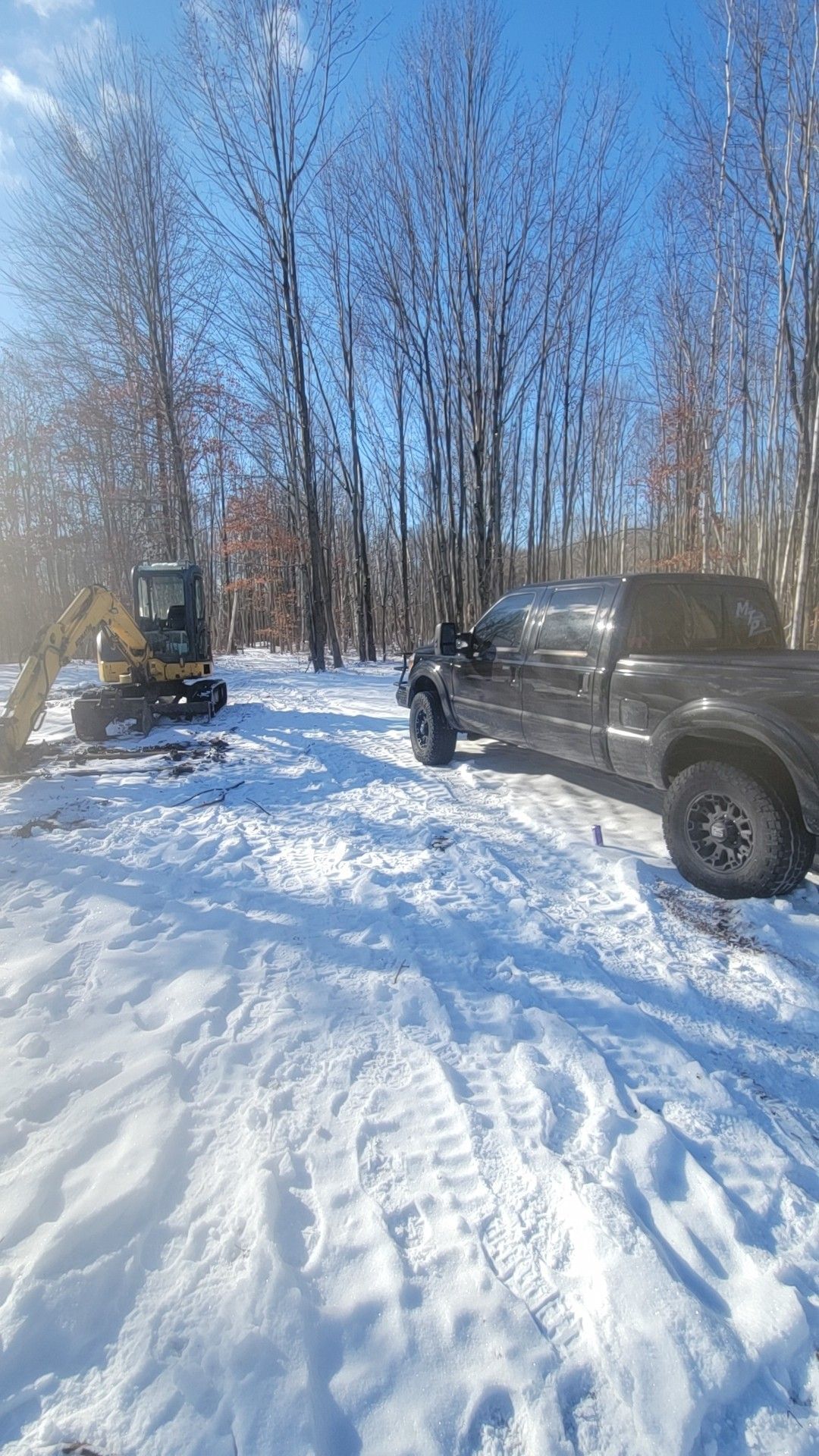 Snowy wooded driveway with a parked dark pickup truck and a small bulldozer in the background.