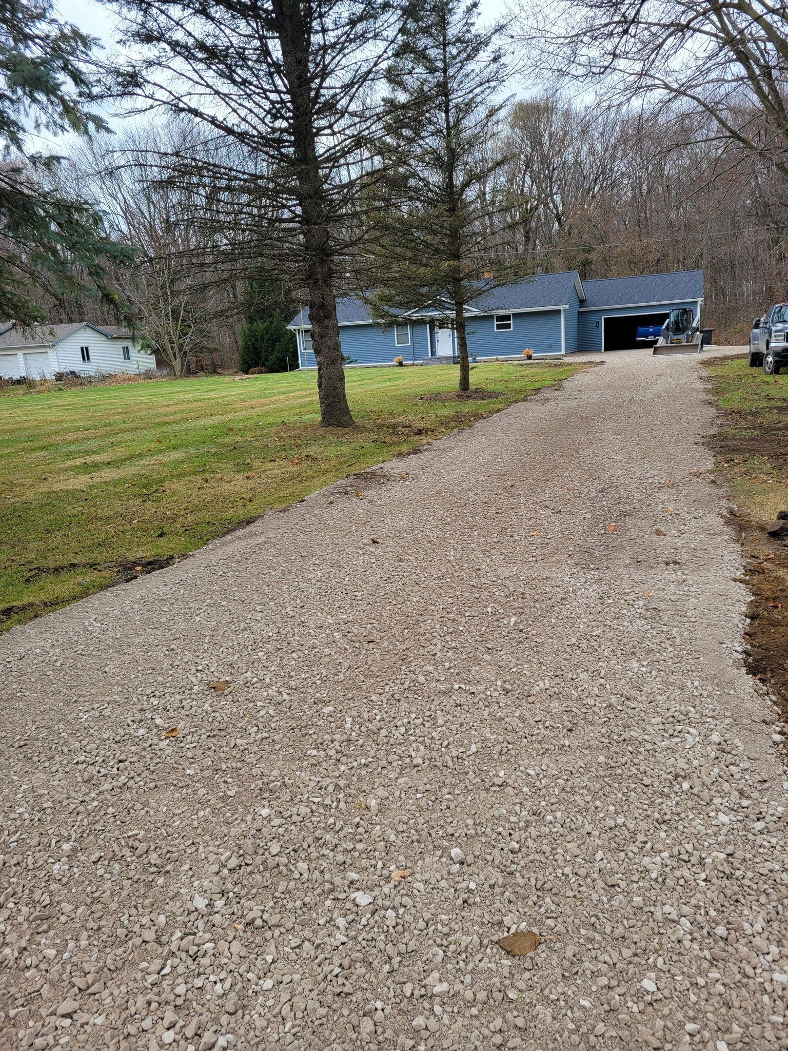 Gravel driveway leading to a blue house with a carport, bordered by leafless trees and winter grass.