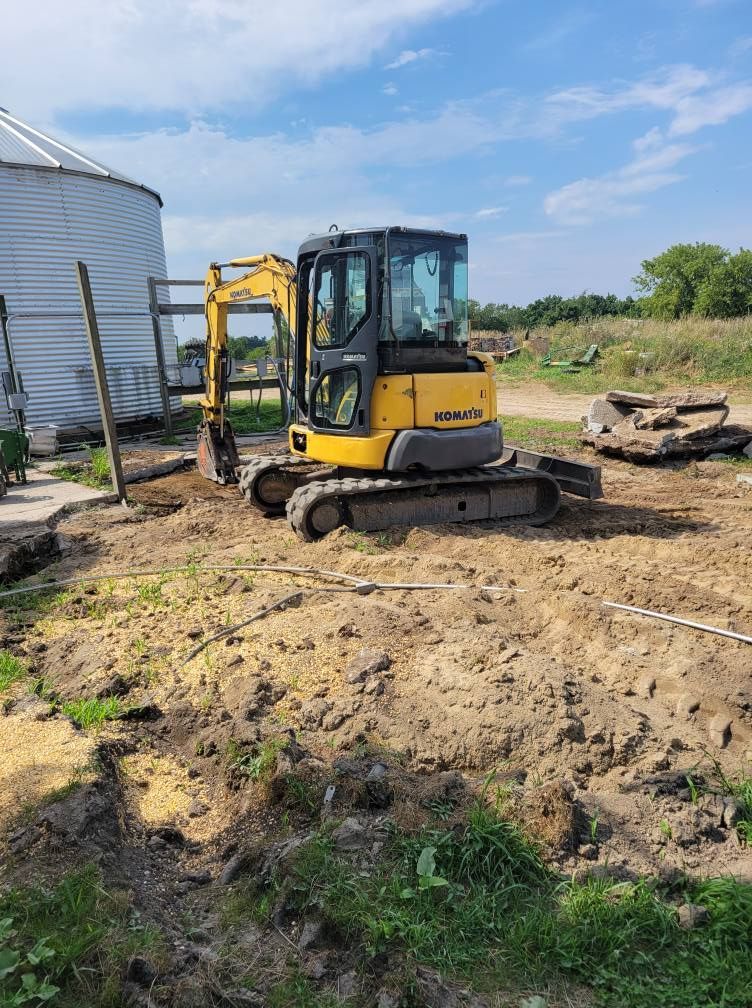 Yellow mini excavator on a dirt lot beside a metal building under a blue sky.