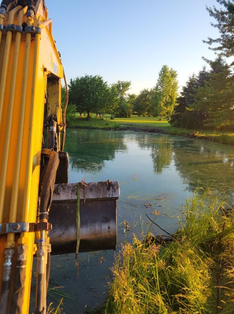 Yellow excavator bucket beside a calm pond with trees reflected in the water at sunset