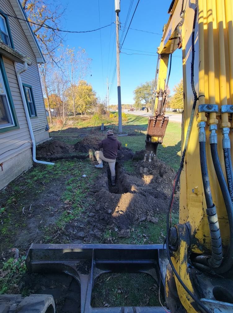 Backhoe trenching beside a house, with a worker standing in the dirt yard.